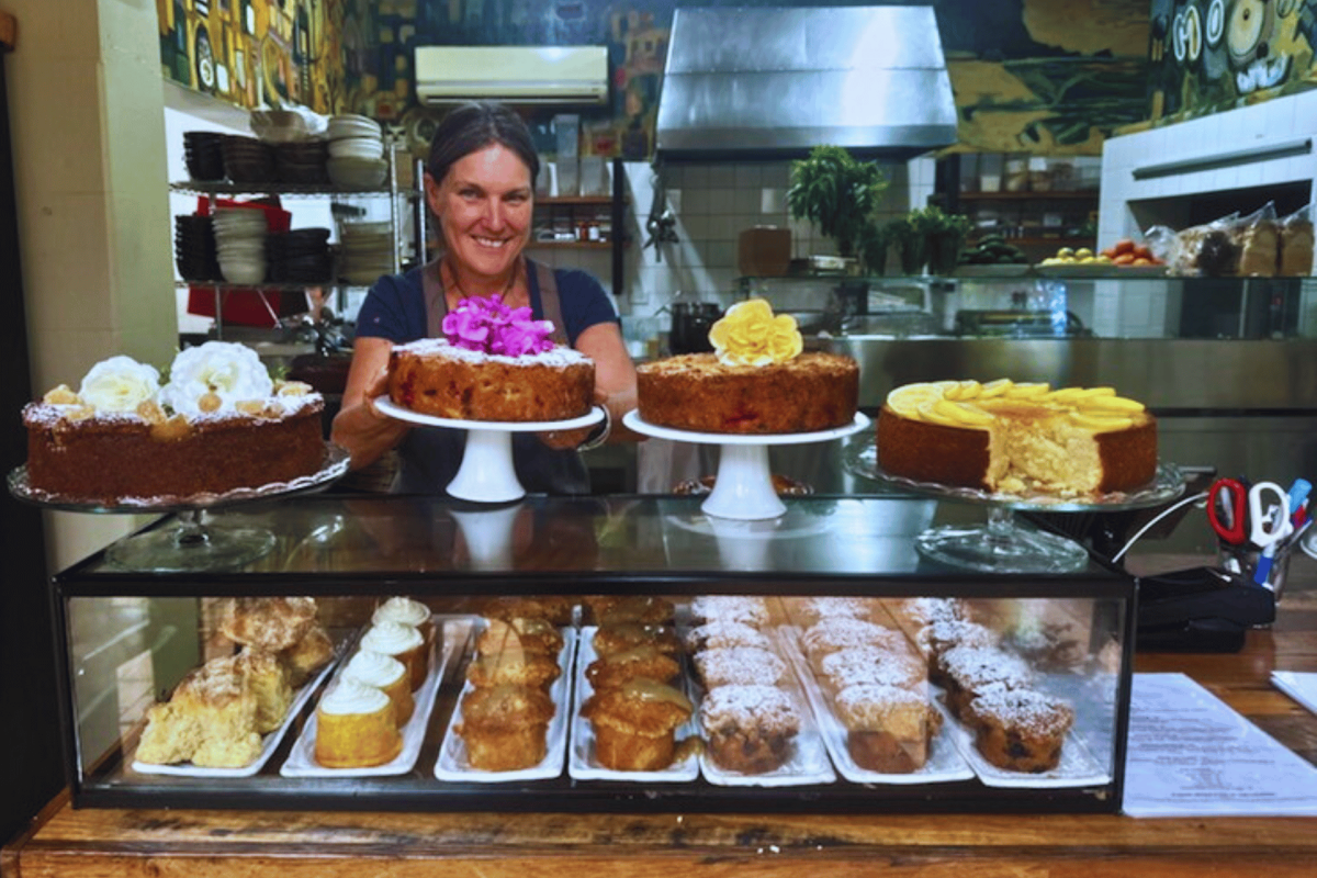 Woman stands behind counter filled with cakes.