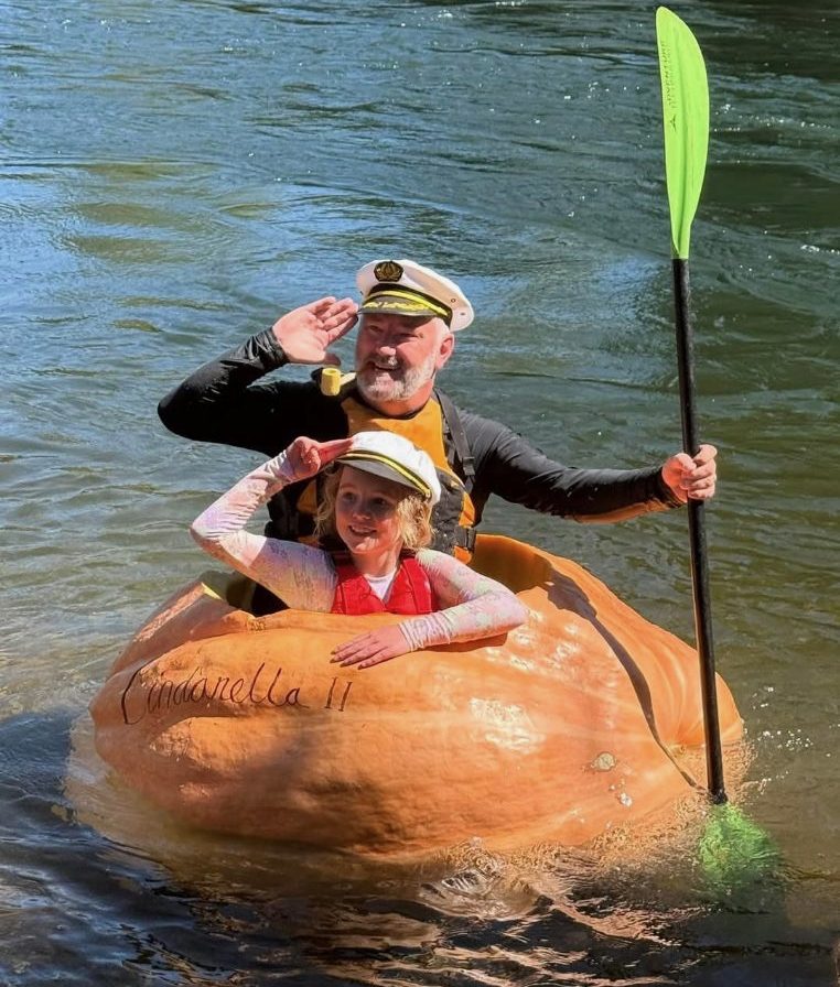 A man and small girl salute from inside a giant pumpkin floating on a river