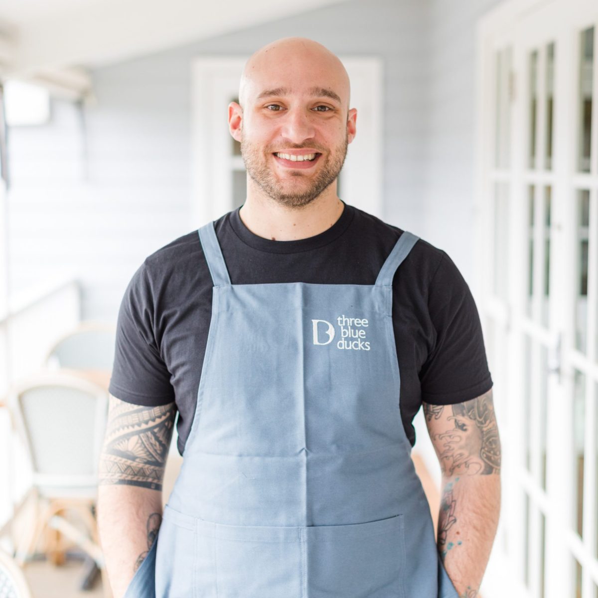 Chef Frankie stands on a white painted verandah wearing a black tshirt and blue apron.