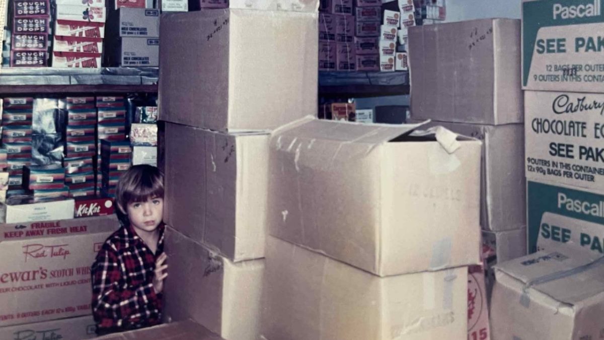 John Rawlinson’s younger brother Matthew dwarfed by cartons containing smaller boxes and bags full of lollies in a small shed the family built when launching their wholesalers business.