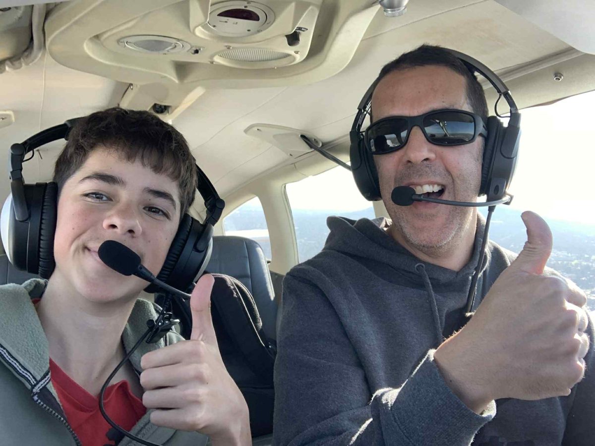 A smiling teenage boy and his father wearing headsets inside a small plane