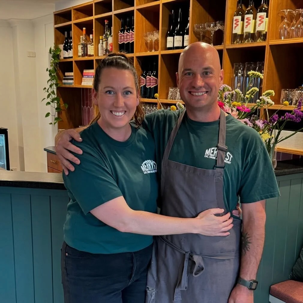 A smiling woman and man in matching Merry St T-shirts stand in front of the bar counter in their restaurant