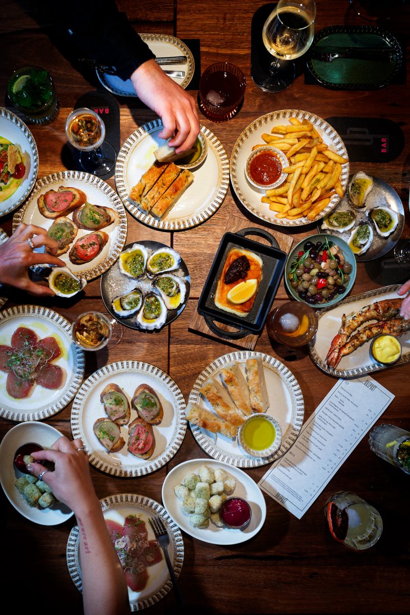 A spread of sharing dishes on a table with arms reaching across to serve.