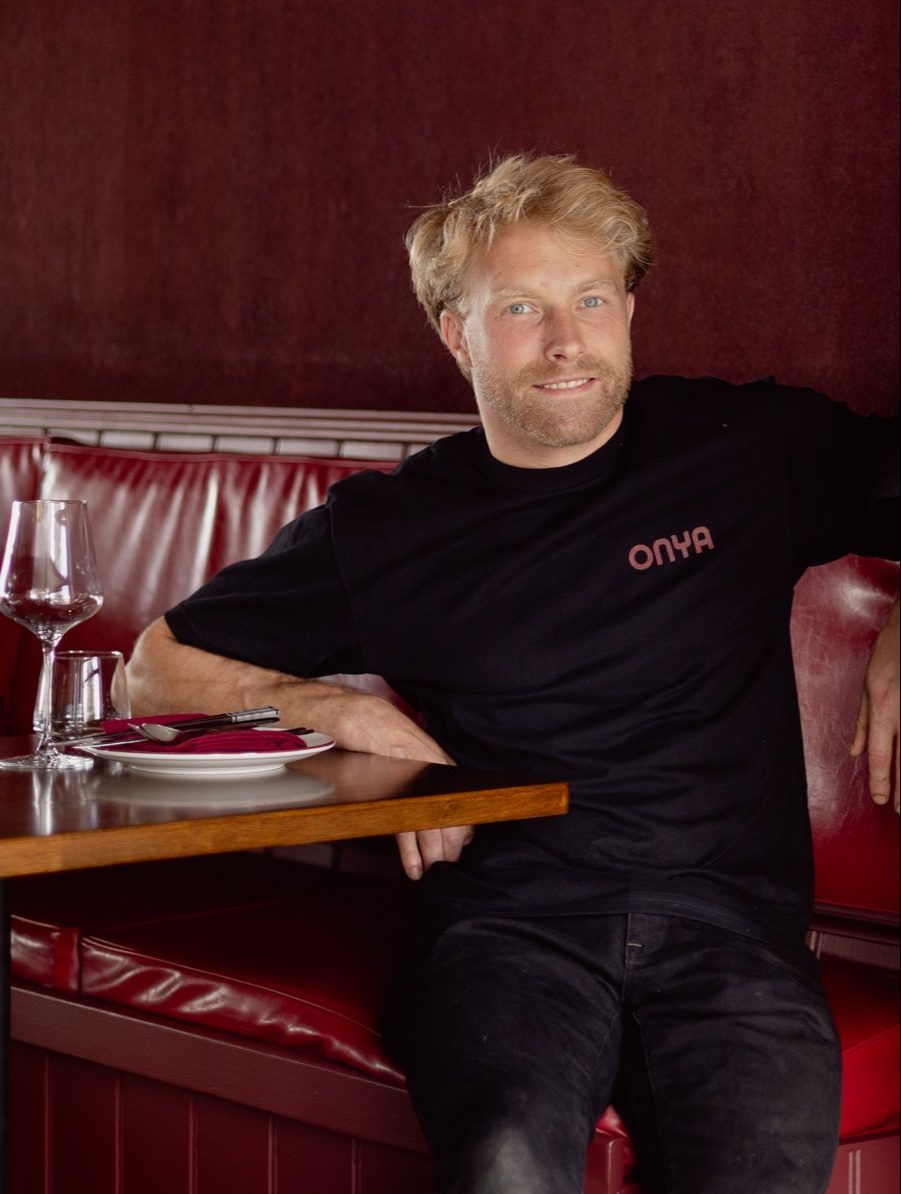 A man sits at a red restaurant booth wearing a black T-shirt which reads Onya in red text.