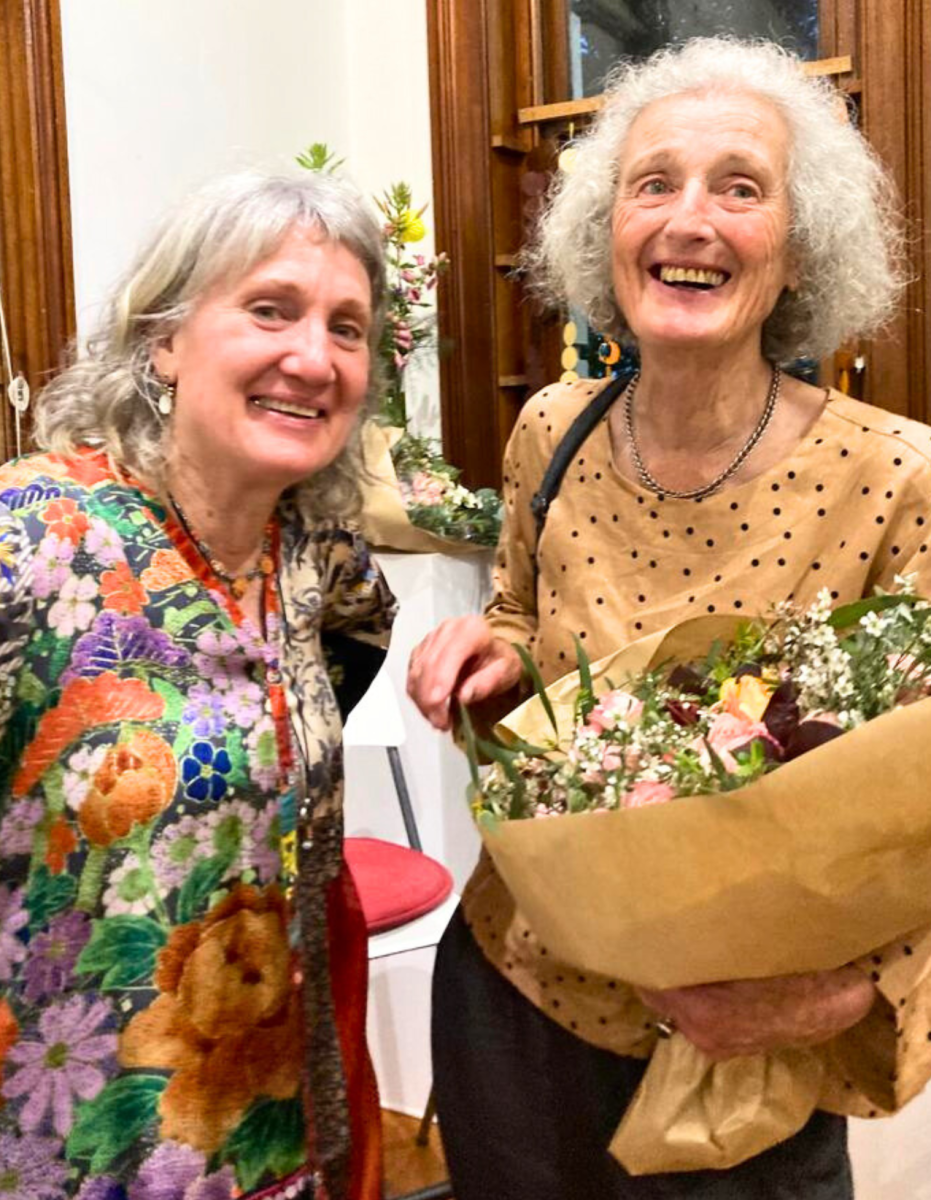 Two women smile at camera, one holds flowers.