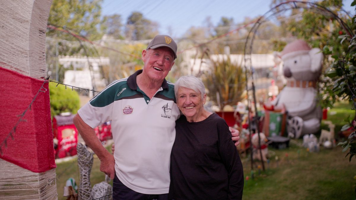 A smiling elderly couple in their Christmas light display, in the daytime