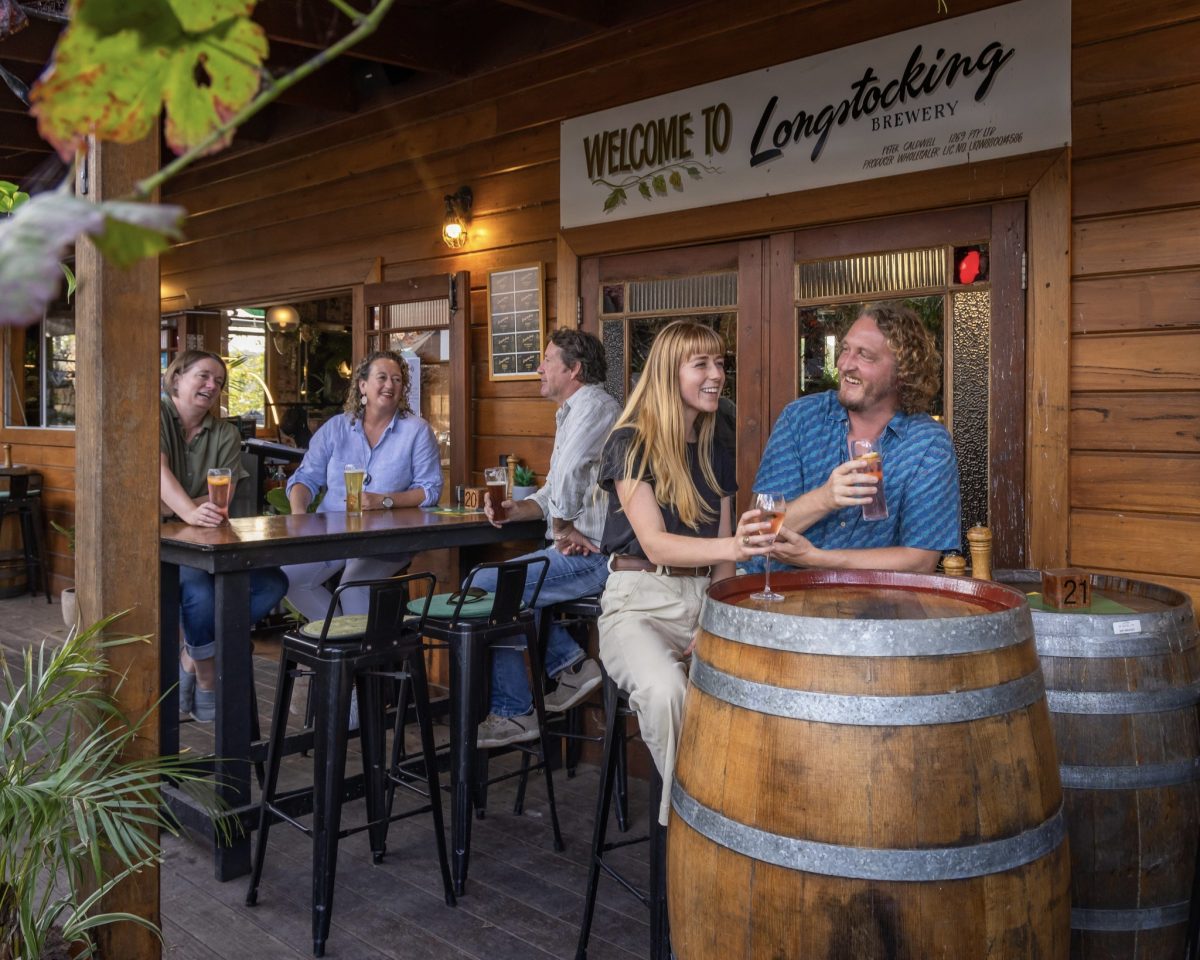People sit at hightop tables and large barrel tables drinking beer outside. A sign reads 'Welcome to Longstocking Brewery'.